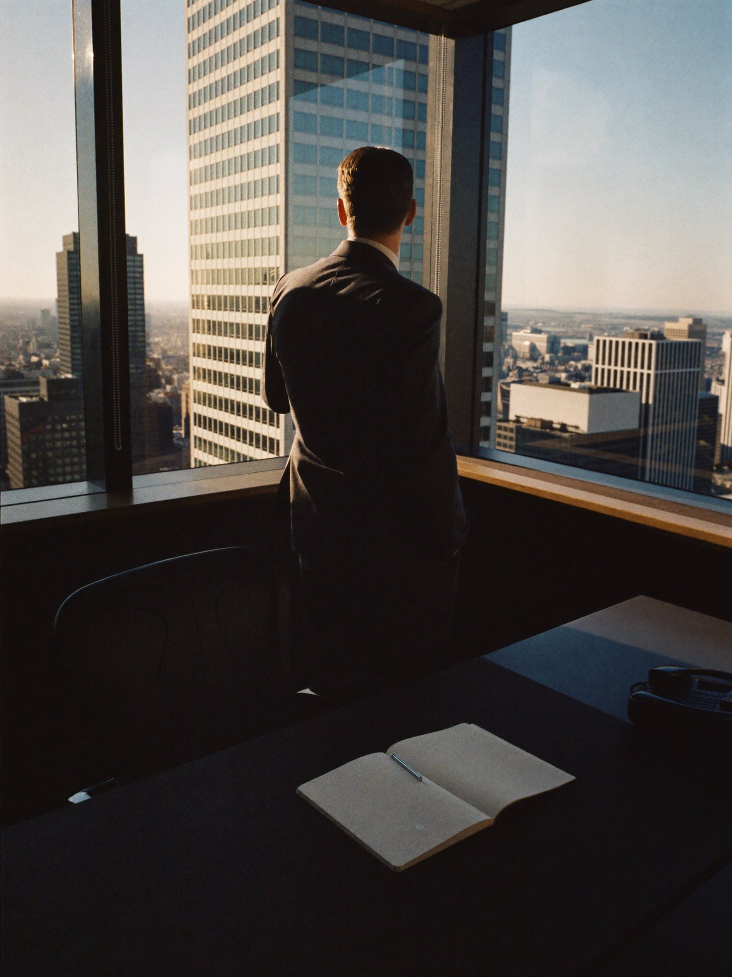 A sales executive at a corner-office window, seen from behind, late afternoon