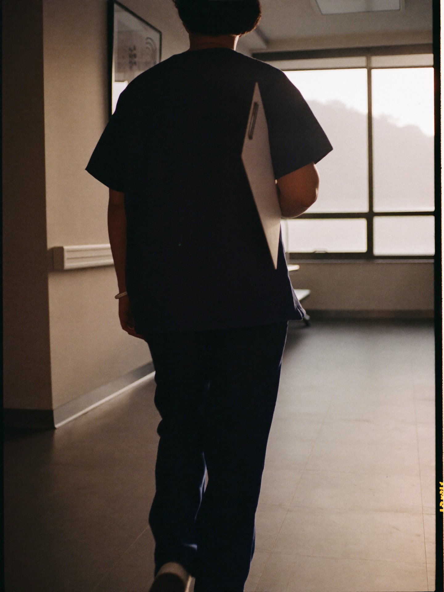 A nurse walking down a quiet hospital corridor toward the morning light